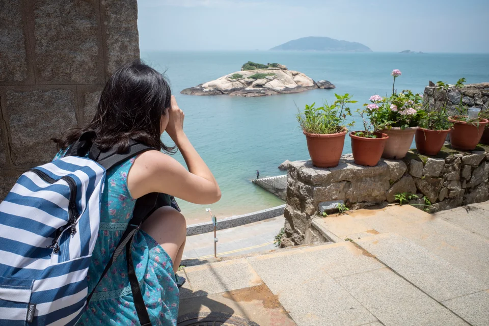 Jeany 背著條紋背包停留在海邊，旁邊的石牆上擺著花盆